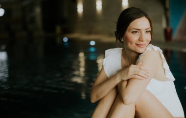 Bir kadın su yansıtan ışıklarla havuzun kenarında oturuyor. Rahatlamış görünüyor ve dizlerine sarılırken hafifçe gülümsüyor. Akşam oldu ve ortam modern..