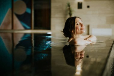 Kadın kapalı bir yüzme havuzunda zaman geçirmekten hoşlanıyor. Gözleri kapalı geri yaslanıyor ve su yakınlardaki sanat eserlerini yansıtıyor. Dekorun modern bir hissi ve sıcacık bir aydınlığı var..