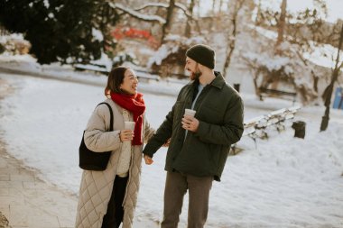 Koreli bir kadın ve beyaz bir erkek, karla kaplı bir parkta el ele tutuşup gülümsüyorlar. Her birinin elinde bir içecek kabı var. Ağaçlar ve banklar karla kaplıdır..