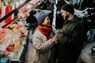 Koreli bir kadın ve beyaz bir erkek kış pazarında neşeli bir anı paylaşıyorlar. Etrafları renkli süslemeler ve ışıklarla çevrili. Ortam canlı ve şenlikli..