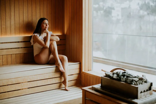 Bir kadın saunadaki ahşap bir bankta sessizce oturuyor. Sıcaklığın tadını çıkarırken rahatlamış görünüyor. Pencerenin dışında, kar zemini kaplıyor, kış olduğunu gösteriyor..