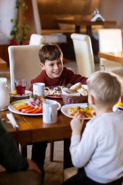 İki genç çocuk çeşitli yiyecek ve içeceklerle dolu bir masada oturuyor. Kahvaltının tadını çıkarırken gülümsemeyi paylaşıyorlar. Masada sebze, ekmek ve meyve var..