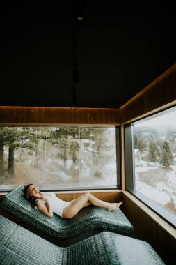 Sessiz bir odada sıcak taştan bir yatakta yatan bir kadın. Güneşli bir günde ağaçların ve karın büyük pencerelerden görülebildiği manzarayı seviyor..