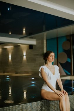 Beyaz mayo giymiş bir kadın kapalı havuzun kenarında sessizce oturur. Ayarlar yumuşak ışıklandırma ve modern dekorasyonu içeriyor. Spa 'nın sakin atmosferinden hoşlanıyor gibi görünüyor..