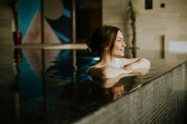 Kolları kenarda dinlenen bir kadın spa havuzunda dinleniyor. Bir anlık rahatlama ve kendine bakmanın tadını çıkarıyor. Sakin ortam modern tasarım elementleri ve sanat eserlerini içerir..