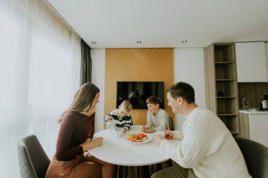 Modern bir mutfakta dört aile üyesi yuvarlak bir masada oturuyor. Çocuklar kahvaltı etmekle meşguller. Güneş ışığı odayı dolduruyor, sıcak bir atmosfer yaratıyor ve yemeklerini paylaşıyorlar..