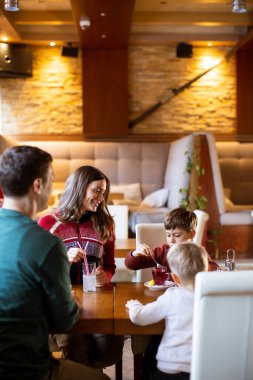 Bir aile restoranda bir masanın etrafında oturur. Ebeveynler konuşurken iki çocuk abur cubur yiyor. Sıcak ışıklar, bu zamanı paylaşırken dostane bir atmosfer yaratıyor..