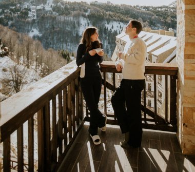 Bir adam ve bir kadın ellerinde içkilerle ahşap bir balkonda durup karlı dağlara bakıyorlar. Güneş sabah gökyüzünde parlıyor, ılık bir atmosfer yaratıyor..