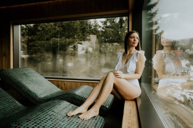 Bir kadın, kaplıcadaki geniş bir pencerenin yanında rahatlar. Tahta bir çıkıntıda otururken ağaçlara bakıyor. Ortam sıcak ve davetkar, rahatlık ve doğaya odaklı..