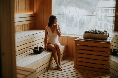 Bir kadın, karla kaplı dağlar ve sıcak buharla çevrili, ahşap bir saunada huzurlu bir anın tadını çıkarır..