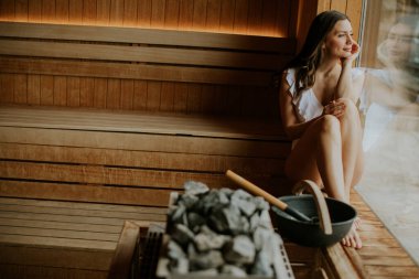 Bir kadın saunadaki ahşap banklarda oturur. Sıcaklığın tadını çıkarırken pencereden dışarı bakıyor. Buhar ve kayalar yakınlarda, rahat bir atmosfer yaratıyorlar..