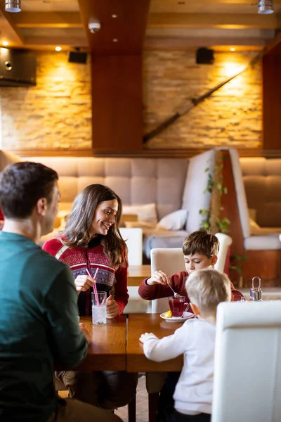 Bir aile restoranda bir masanın etrafında oturur. Ebeveynler konuşurken iki çocuk abur cubur yiyor. Sıcak ışıklar, bu zamanı paylaşırken dostane bir atmosfer yaratıyor..