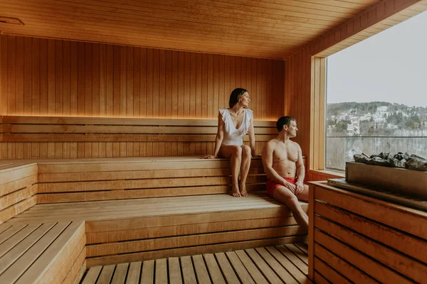 İki insan saunada eğleniyor. Bir kişi bankta otururken diğeri duvara yaslanıyor. Binanın dışını gösteren bir pencere.