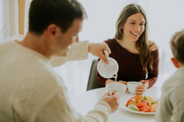 Bir adam fincana çay dökerken bir kadın ona gülümsüyor. Genç bir çocuk masanın üzerindeki renkli bir meyve tabağına bakıyor. Sabahleyin parlak bir mutfaktalar..
