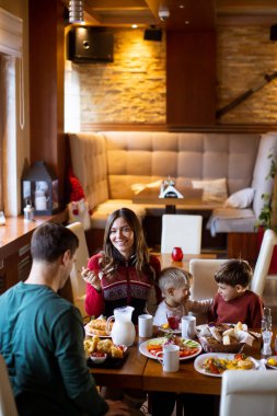 Bir kadın, neşeli çocukları ve eşiyle brunch 'tan keyif alır. Sıcak bir kafe atmosferinde gülümsemelerini ve lezzetli yemeklerini paylaşır..