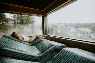 Bir kadın, sakin bir kaplıcadaki sıcak bir taş yatakta huzur içinde, dışarıdaki büyüleyici dağ manzarasının tadını çıkarıyor..