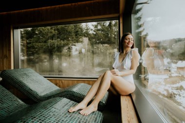 Bir kadın, kaplıcada pencerenin kenarında oturur ve dışarıdaki karla kaplı ağaçların tadını çıkarır. Bir mayo giyer ve şezlongda dinlenirken mutlu bir şekilde gülümser..