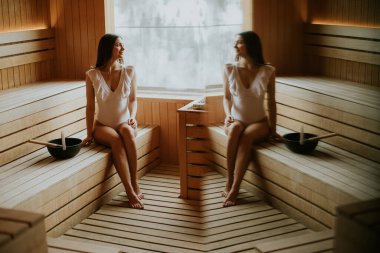 Sıcak bir saunada huzur içinde oturan bir kadın huzurlu atmosferin tadını çıkarıyor ve kendini yenilenmiş hissediyor. Ahşap döşeme onun deneyimine sıcaklık katıyor..