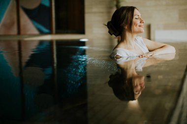 Bir kadın kapalı bir kaplıcadaki havuzda dinleniyor. Gözleri kapalı oturur ve nazikçe gülümser. Su onun imajını ve etrafındaki huzurlu çevreyi yansıtıyor..