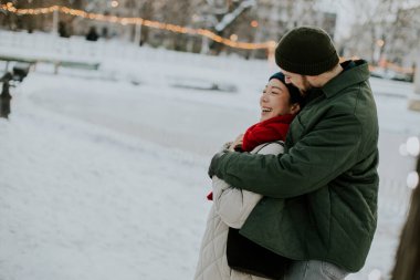 Koreli bir kadın ve beyaz bir erkek akşam karlı bir parkta sıcak bir kucaklaşmayı paylaşıyorlar. Işıklar ve kış atmosferiyle çevrili..