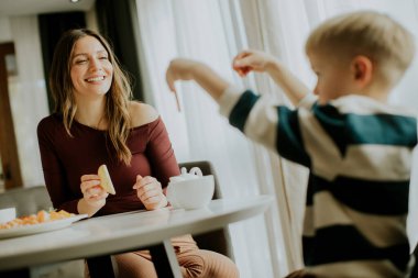 Bir kadın bir masada oturur, bir çocukla atıştırmalık paylaşırken gülümser. Çocuk yemekle uzanırken, parlak bir odadaki masada bir tabak abur cubur oturuyor..