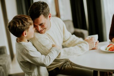 Bir baba ve oğlu, parlak doğal ışığın olduğu sıcak bir odada, bir masada birlikte oturuyorlar. Masada sıcak aile bağlarını gösteren yiyecek ve içecekler varken keyifli bir anı paylaşıyorlar..