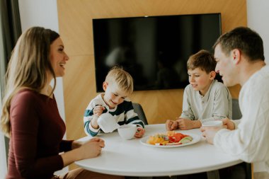 Bir aile kahvaltı için bir masanın etrafında toplanır. İki erkek çocuk ebeveynlerinin yanında oturuyorlar, yiyecek ve içeceklerini paylaşıyorlar. Gülümsüyorlar, konuşuyorlar, birlikte mutfaktan keyif alıyorlar..
