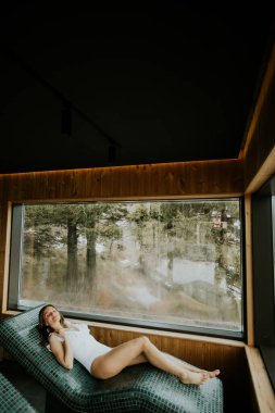 Bir kadın büyük bir pencerenin yanındaki sıcak bir taş yatakta dinlenir. Ağaç ve doğa manzarasıyla baş başa vakit geçirmeyi seviyor. Güneş ışığı pencereden girer ve sıcak bir alan yaratır..