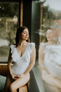 Beyaz mayo giymiş bir kadın büyük bir pencerenin yanında poz veriyor. Düşüncelerini yansıtırken sıcak güneş ışığından hoşlanır. Dağlar arka planda görünür..