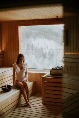 Sıcak, ahşap duvarlar saunada sessizce oturan bir kadını sarar, sakin, karlı manzaraya bakar..