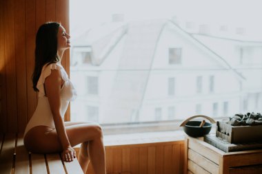 Bir kadın, pencerenin yanındaki saunada vakit geçirmekten zevk alır. Tahta bir bankta mayoyla oturuyor. Güneş ışığı camdan yansıyor, dışarıdaki binaları aydınlatıyor..