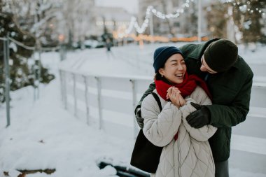 Koreli bir kadın ve beyaz bir adam karlı bir parkta birlikte gülüyorlar. Sıcak bir şekilde giyinmiş ve arka planda tatil ışıklarıyla dışarıda vakit geçiriyorlar. Kar zemini kaplıyor..
