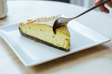 Bir insan beyaz tabakta bir dilim peynirli kek kesmek için kaşık kullanır. Tatlı yeşil bir katman içeriyor ve doğal ışıkla gelen bir kafe ortamında oturuyor..