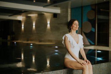 Bir kadın kapalı bir alanda havuzun kenarında oturuyor. O bir mayo giyer ve su kenarında dinlenirken gözlerini kapatır. Yumuşak ışıklar etrafındaki boşluğu aydınlatıyor..