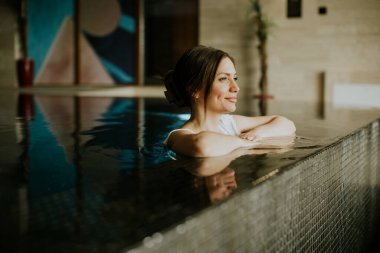 Bir kadın şık bir kapalı havuzda suyun tadını çıkartıyor. Kollarını kenarda dinlendiriyor ve memnun görünüyor. Doğal ışık boşluğu doldurur ve rahatlatıcı bir hava yaratır..