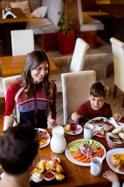 A family gathers around a table to share breakfast in a restaurant. The adults smile as the children eat fruits and pastries, enjoying a meal filled with various dishes.