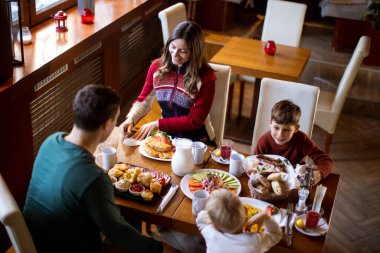 A family of four shares breakfast at a restaurant. They sit around a table filled with food, drinks, and fruit. Children watch as their parents serve food to each other.
