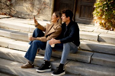 İki arkadaş binanın dışındaki taş basamaklarda oturuyor. Ellerinde içkiler var ve gülümserken birbirlerinin fotoğraflarını çekiyorlar. Güneş üzerlerine parlıyor, sıcak bir atmosfer yaratıyor..