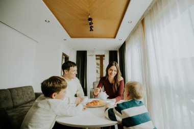 Yuvarlak bir masada yemek yiyen bir aile. İki oğlan izlerken bir kadın çay dolduruyor. Bir adam gülümser ve sohbet eder. Oda aydınlık ve büyük pencereleri var..