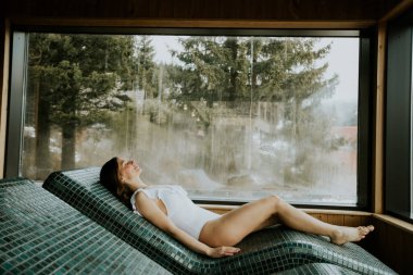 Sıcak taştan bir yatakta uzanan bir kadın. Büyük bir pencereden içeri güneş ışığı giriyor. Rahat görünüyor ve dışarıdaki ağaçların manzarasından hoşlanıyor. Olay yeri spa ya da sağlık inzivasına çekilmeyi öneriyor..