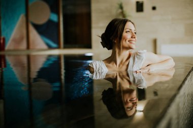 Bir kadın havuzda oturur, kenara yaslanırken gülümser. Arka planda parlak duvarlar ve modern dekorlar görülebilir. Sahne öğleden sonra bir kaplıcada geçiyor..