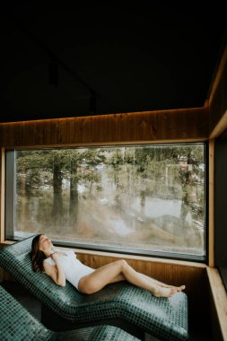 Bir kadın bir odanın içindeki sıcak taş bir yatakta yatıyor. Dışarıdaki ağaçları ve doğayı gösteren büyük bir pencerenin yanında. Sıcak ve aydınlık bir gün ve o rahat görünüyor..