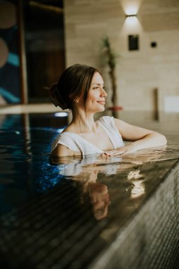 Kaplıcadaki kapalı havuzda rahatlayan bir kadın. Sıcak suyun tadını çıkarırken kenara yaslanıyor. Dekorun yumuşak ışıkları ve rahatlatıcı bir atmosferi var. Rahatlamak için mükemmel..