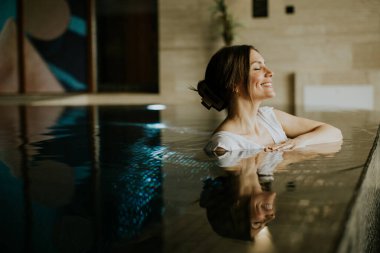 Bir kadın sağlık merkezindeki havuzda dinleniyor. Mutlu bir gülüşü var ve kenarına yaslanıyor. Dekorun pürüzsüz yüzeyleri ve yumuşak ışıklandırması var. Bitkiler arka planda görülebilir.