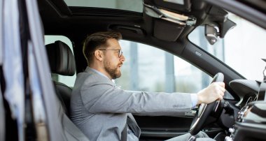 Takım elbiseli bir adam lüks bir arabanın sürücü koltuğunda oturuyor. Bir şehirde araba sürerken odaklanmış görünüyor. Güneş parlıyor ve pencerelerden yansıyor. Modern bir bina görülebilir..