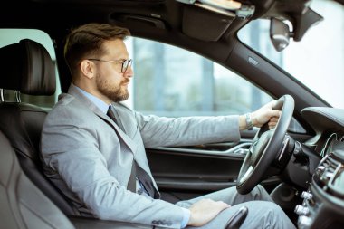 Takım elbiseli bir adam arabanın içinde oturur ve modern bir şehrin içinden geçer. Direksiyonu tutarken odaklanmış görünüyor. Güneş pencerelerden süzülüyor.