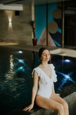 Havuzun yanında mayo giymiş bir kadın oturuyor. Kapalı gözlerle anın tadını çıkarıyor. Kapalı alanda pürüzsüz duvarlar ve arka planda renkli sanatlar var..