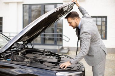 Takım elbiseli bir adam şehir merkezine park ederken arabanın kaputunun altına bakar. Güneş tepede parlarken ve arka planda binalar varken motoru yakından kontrol ediyor..