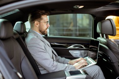 Resmi kıyafetler giymiş bir adam arabanın arka koltuğunda oturur. Seyahatteyken dizüstü bilgisayarına odaklanıyor. Sahne araba pencerelerinin dışında şehir manzarasını gösteriyor..