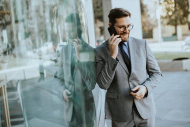 Takım elbiseli bir adam ofis binasının önünde duruyor. Telefonla konuşurken gülümsüyor. Cam duvar onun görüntüsünü yansıtıyor. Arka planda insanlar ve arabalar bulanık.